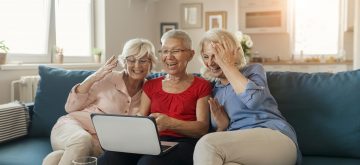 Three lovely senior mature women at home using a laptop in the living room sharing good time together while  looking at a laptop computer and laughing at something on the screen. Smiling senior friends looking at laptop together at home on couch